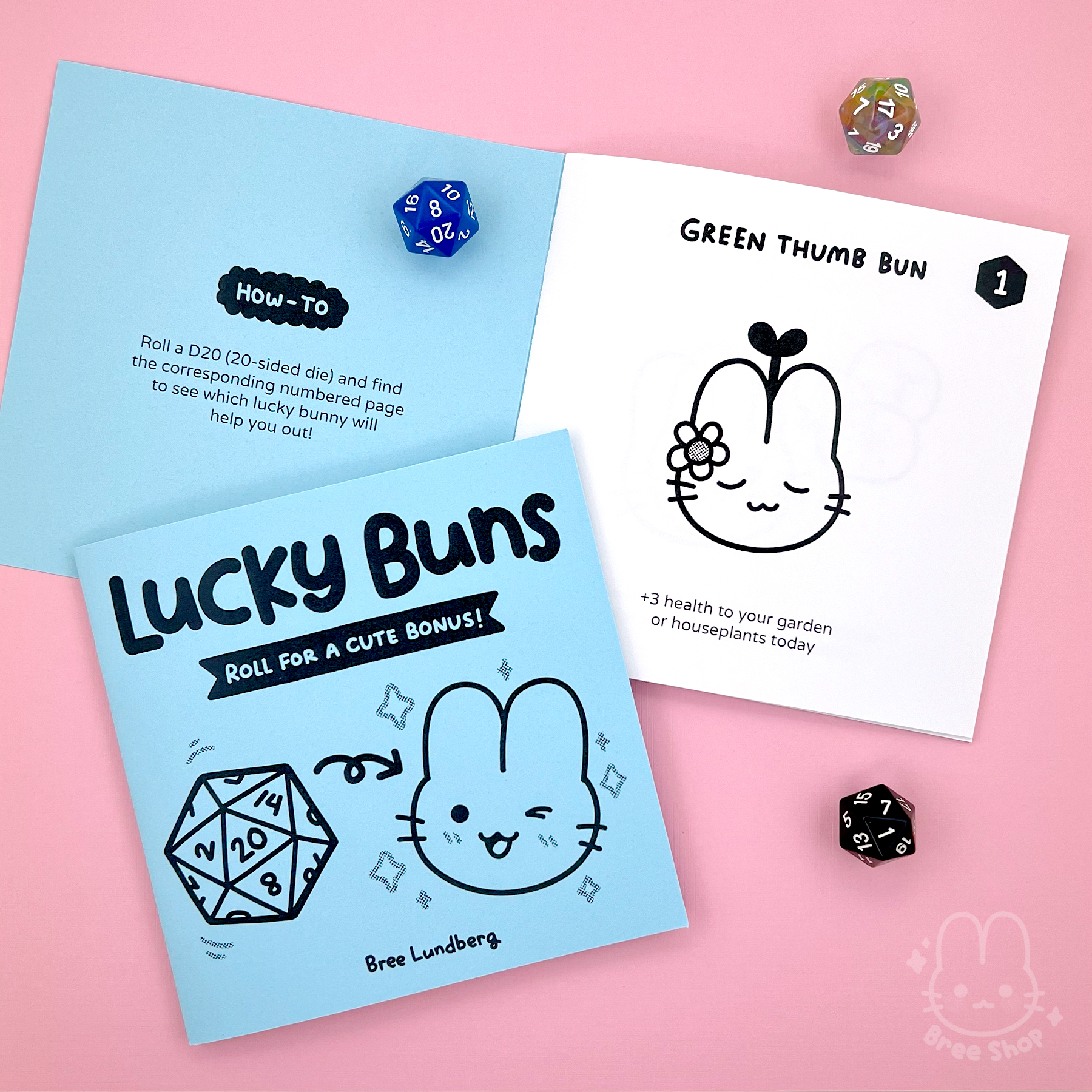 Photo of a pastel square blue zine that says "Lucky Buns, Roll for a Cute Bonus" on the cover with an illustrated D20 and a cute bunny face. Another copy is laying opened showing a "how-to" page and cute black and white art inside.