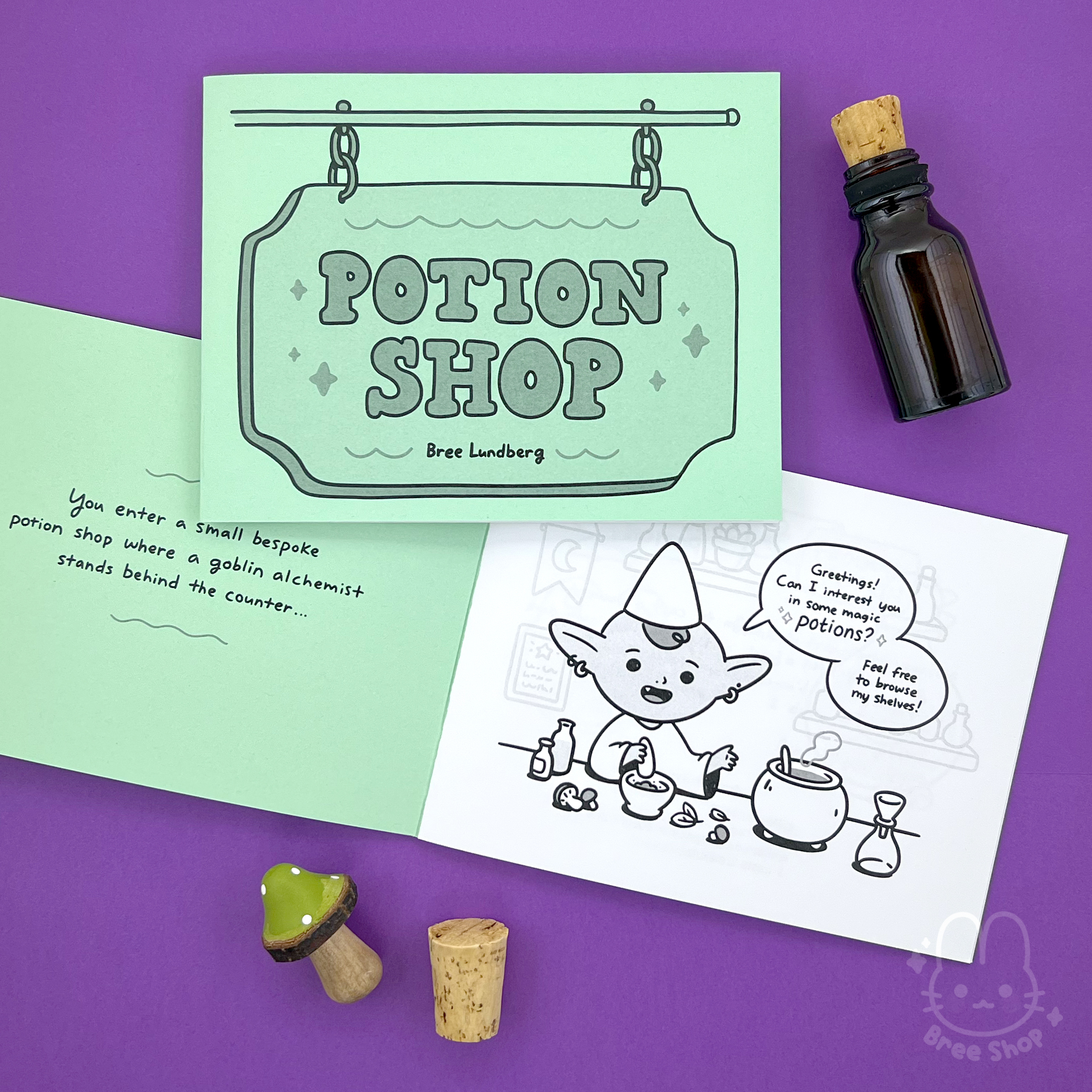 Photo of a pastel green zine that says "Potion Shop" on the cover with an illustrated shop sign, it's laying on another copy that is opened and showing a cute illustrated goblin character preparing potions with speech bubbles welcoming you to the shop.