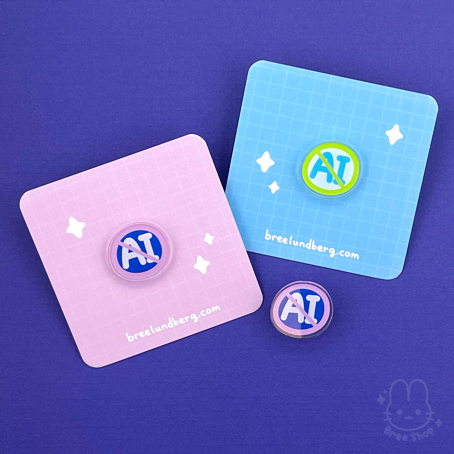 Photo of round-ish acrylic pin badges on backing cards. The pins are circles with the letters "AI" crossed out. There are two color versions. One is pink and dark blue, the other is green and light blue.