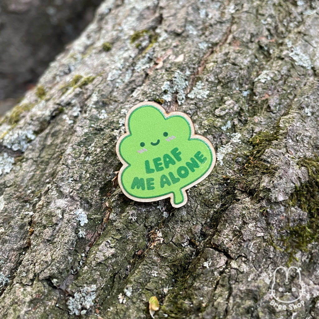 Photo of wooden pin with a cute design of a smiling green leaf and the words "leaf me alone" on it. The pin is laying outside on tree bark.