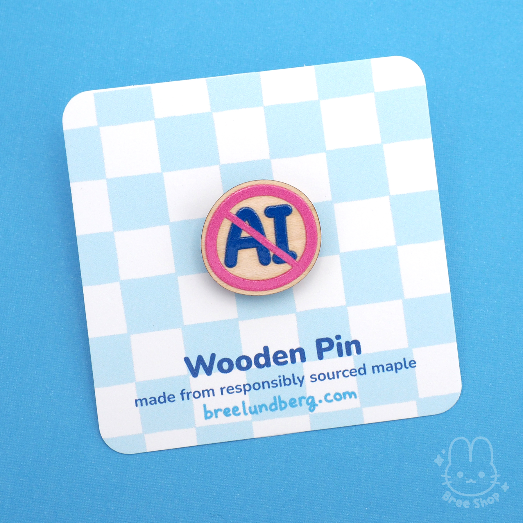 Photo of round-ish wood pin with blue letters "AI" in a crossed out pink circle. It's attached to a backing card with light blue checker pattern and info about the pin.